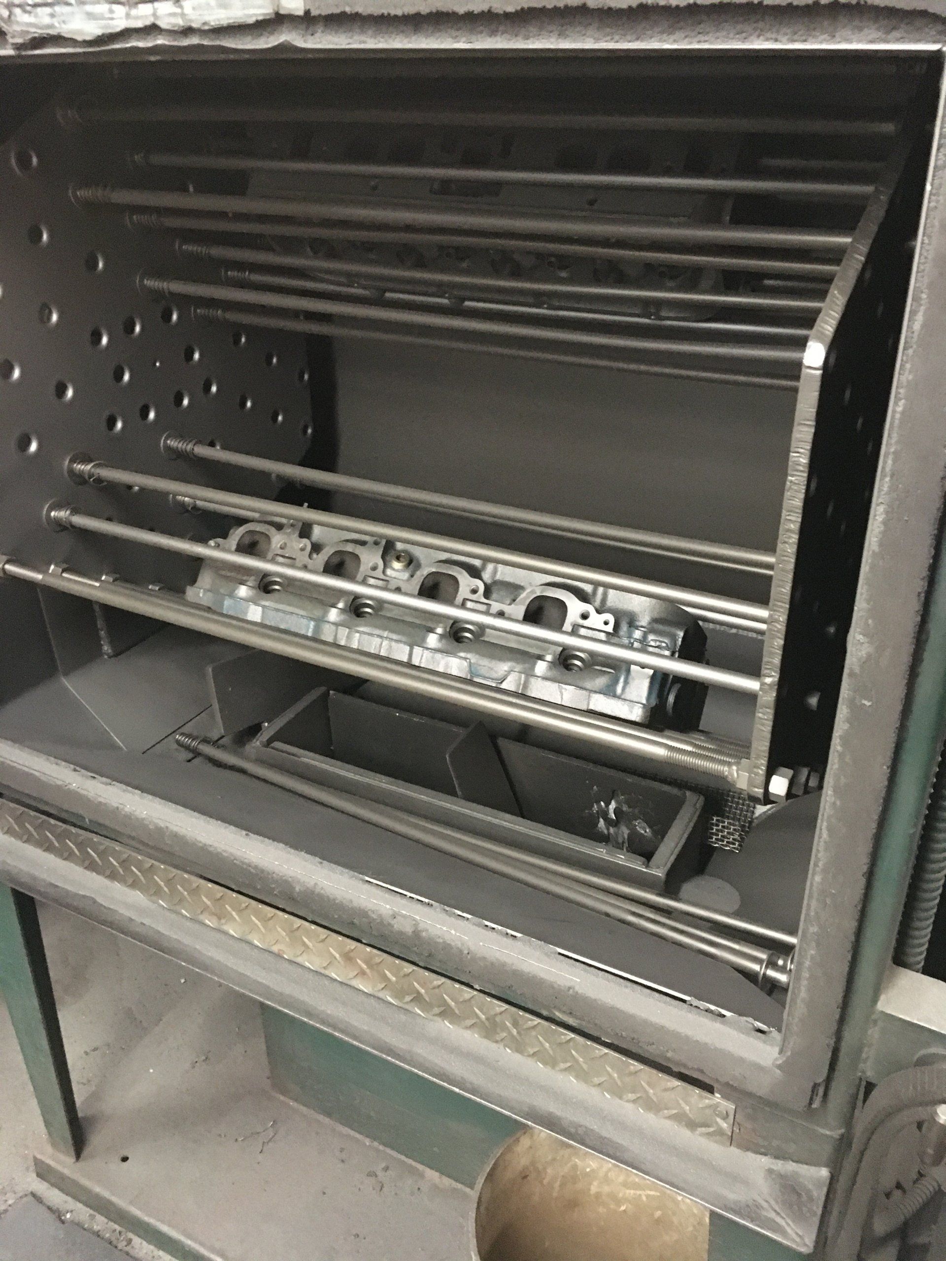 Engine cylinder head inside an industrial oven with metal racks.