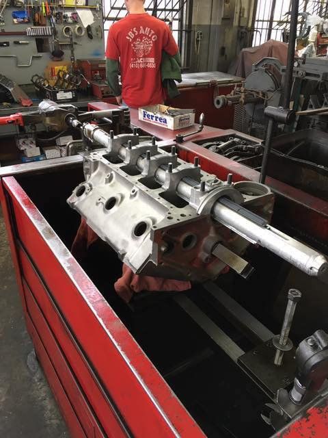 Engine block being worked on in a machine shop. A person in a red shirt stands near the machinery.