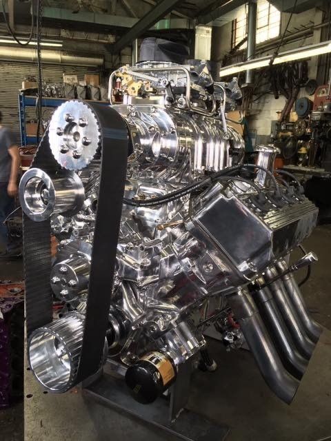 Shiny, polished V8 engine with a large belt and headers, in a workshop setting.