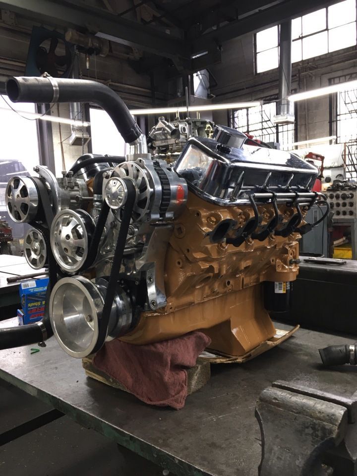 Gold-painted engine with chrome accents, pulleys, and carburetors sits on a workbench in a workshop setting.