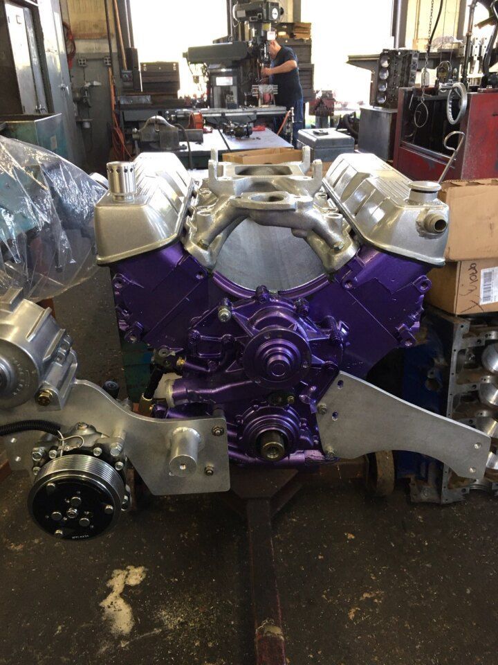Purple engine block with silver components in a workshop. A person is working in the background.