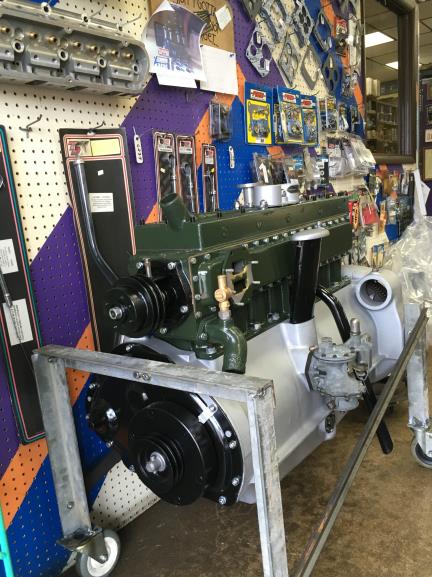 Green and silver vintage car engine displayed on a stand in a shop, with parts and tools in the background.