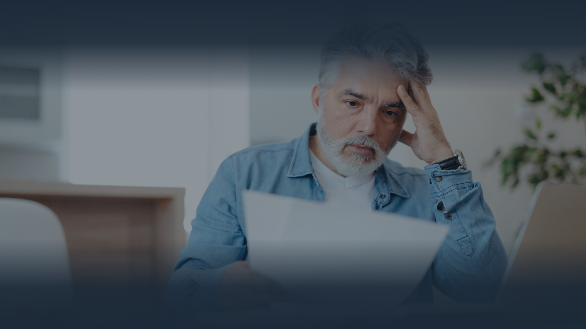 Man looking at paper, hand on head, concerned expression. Indoors.