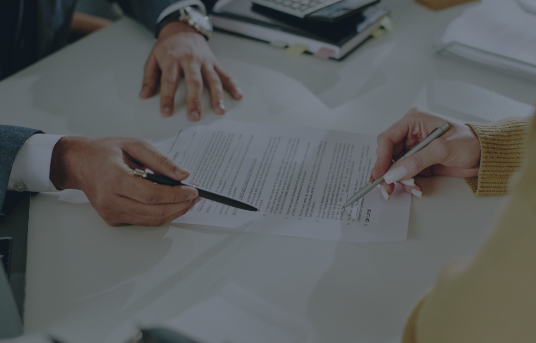 Two people shaking hands over a desk with a laptop and documents near a window.