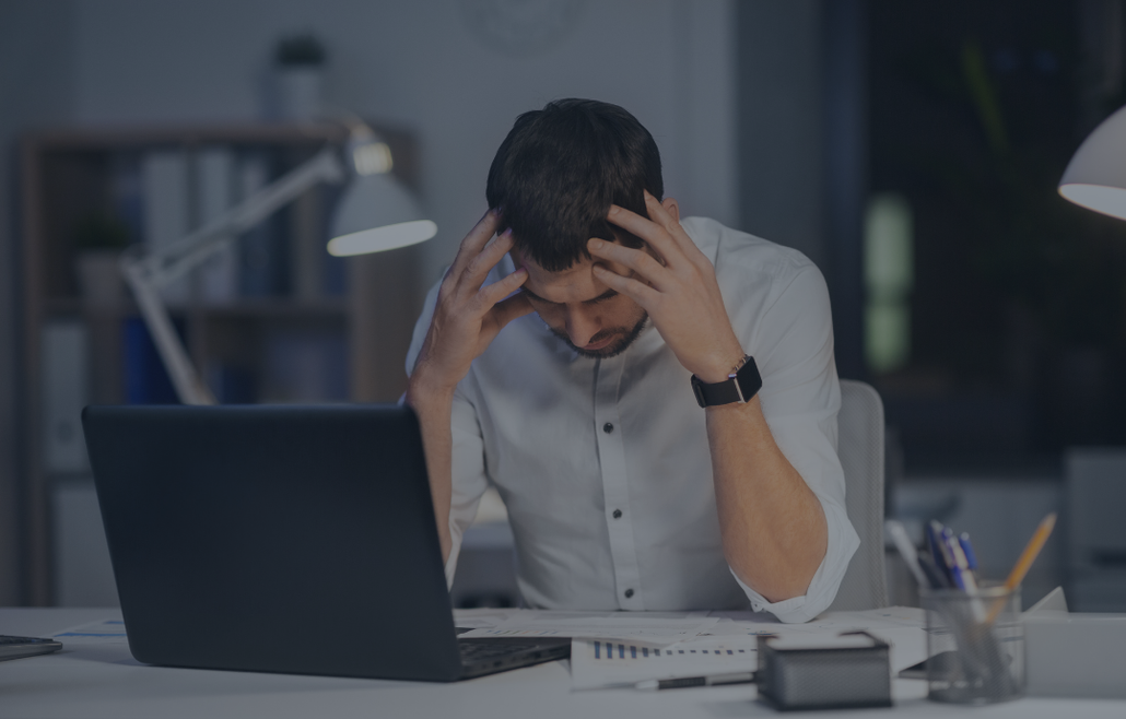 Man stressed at a desk, head in hands, laptop open, paperwork, dimly lit office.