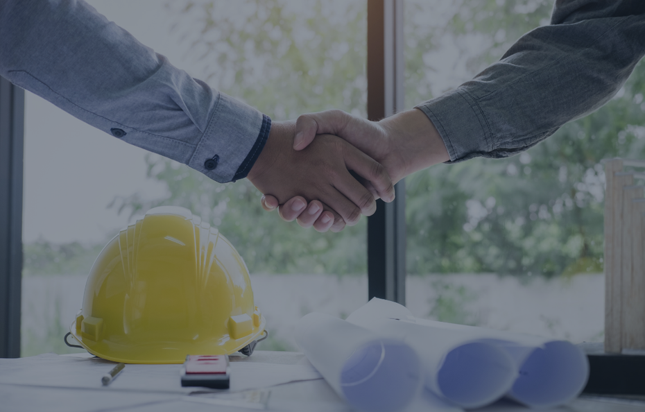 Two people shaking hands over a table with a yellow hard hat and blueprints.