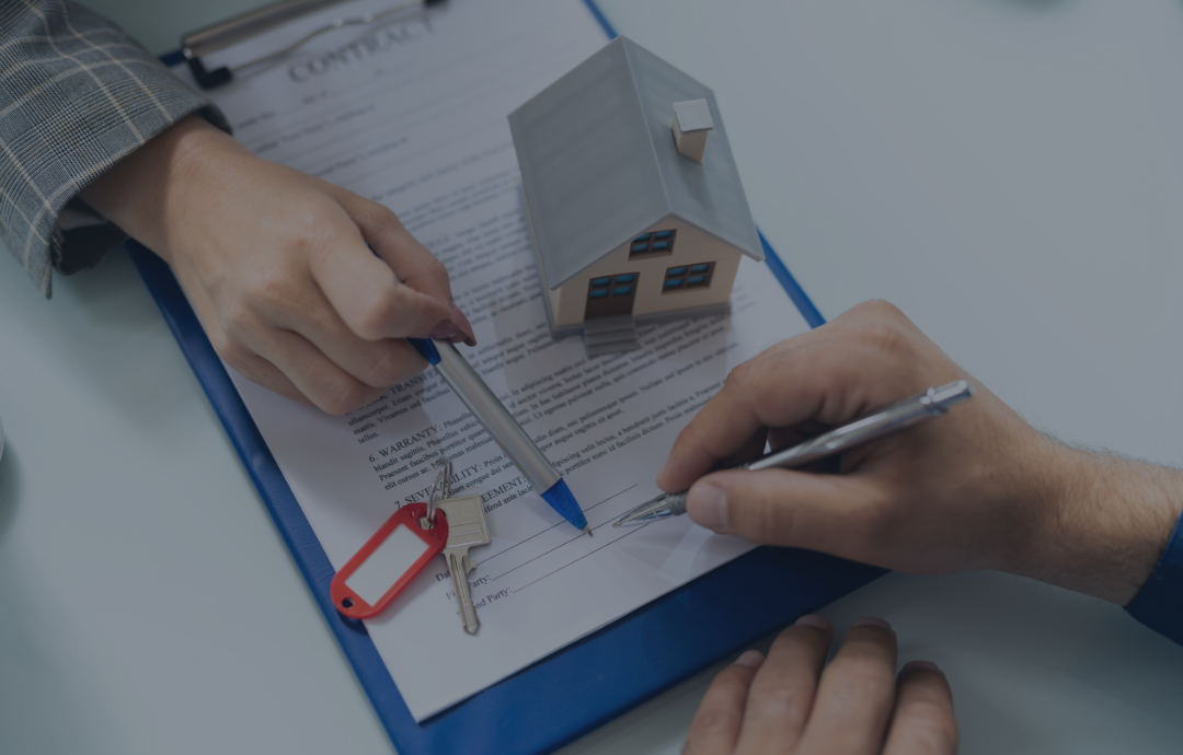 Hands signing a real estate contract with a miniature house and keys on the table.