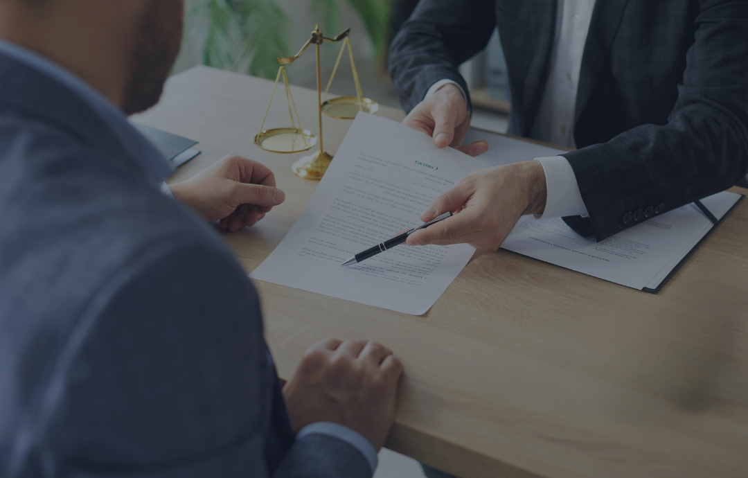Two people shaking hands over a desk with a laptop and documents near a window.
