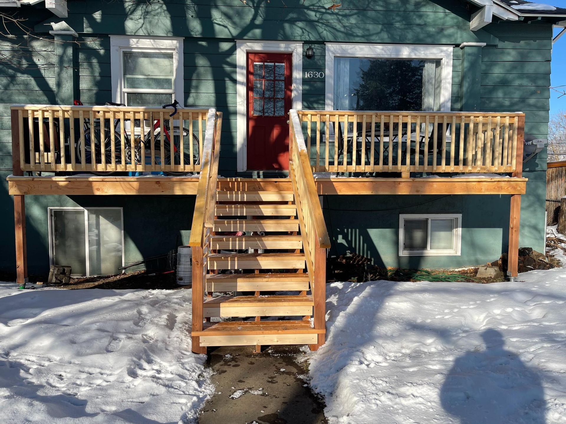 Wooden deck with stairs leading up to a red door. House is painted teal with snow on the ground.