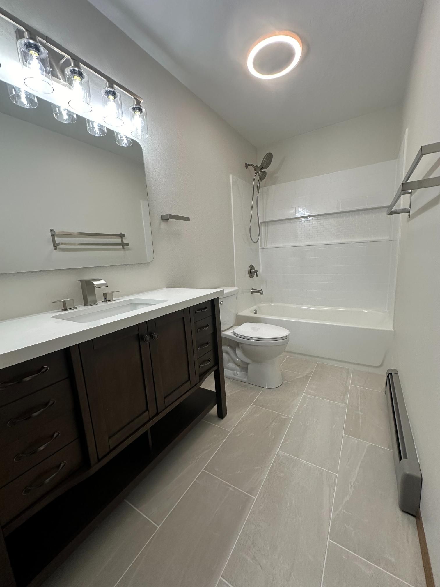 Bathroom with dark wood vanity, white countertop, and gray tiled floor. White tub/shower combo.