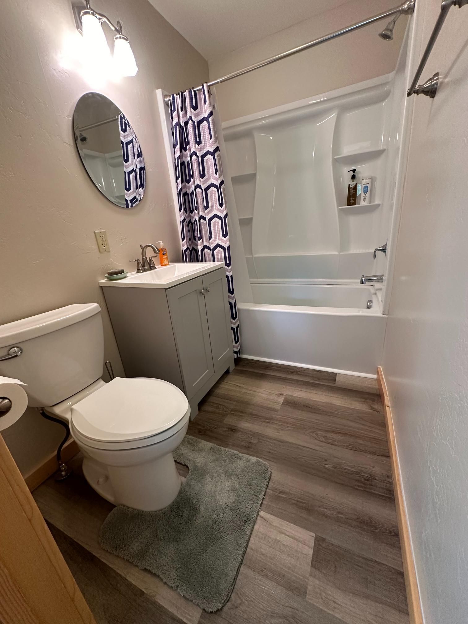Bathroom with gray vanity, toilet, bathtub, shower curtain, oval mirror, and wooden floor.