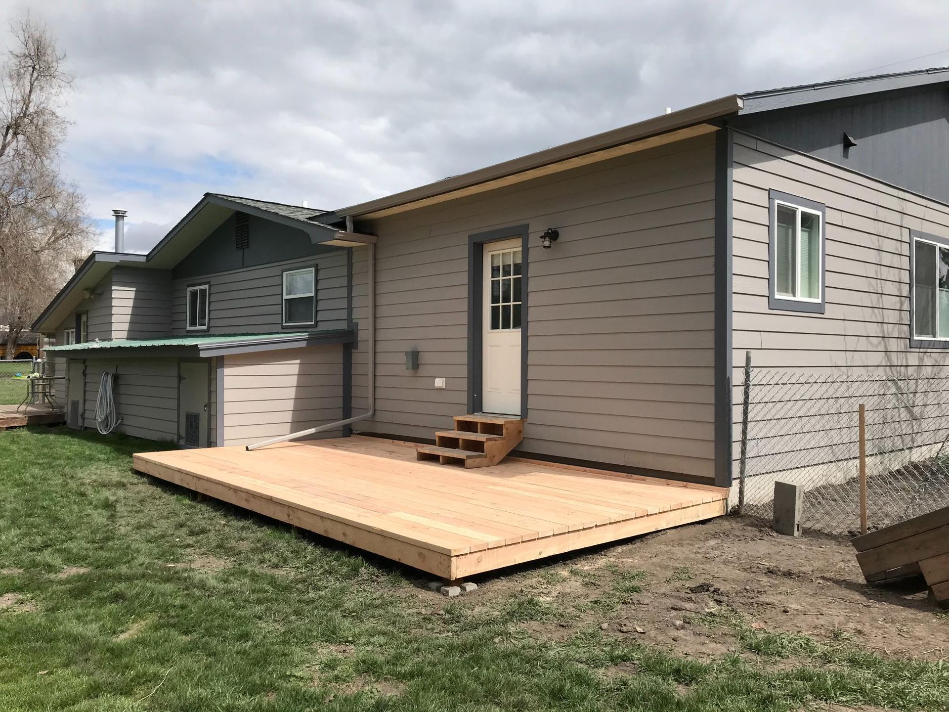 Wooden deck attached to a gray house with a white door. Green grass surrounds the deck.