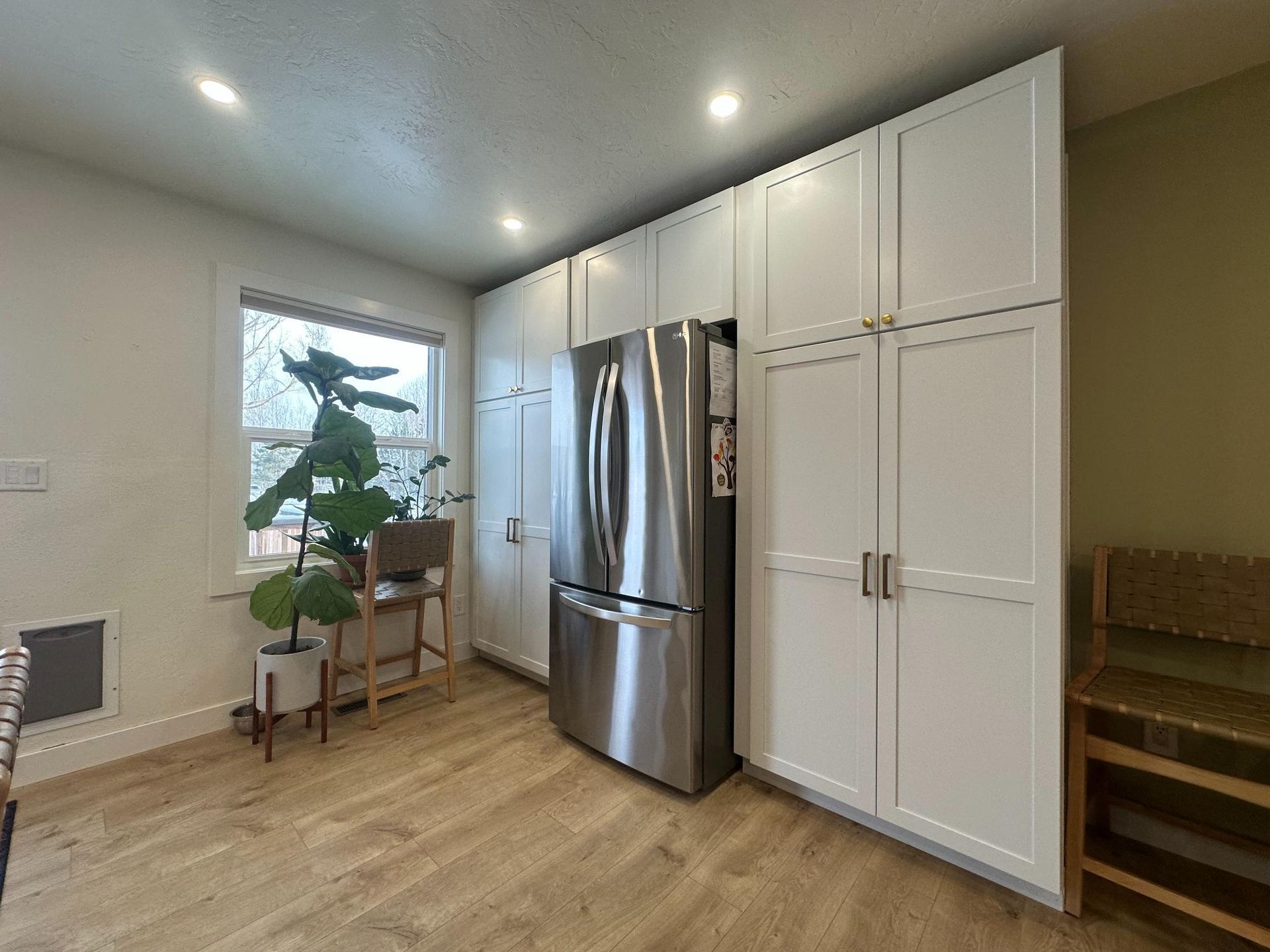 Kitchen with white cabinets, stainless steel fridge, and wooden floors. A window and plants are also visible.