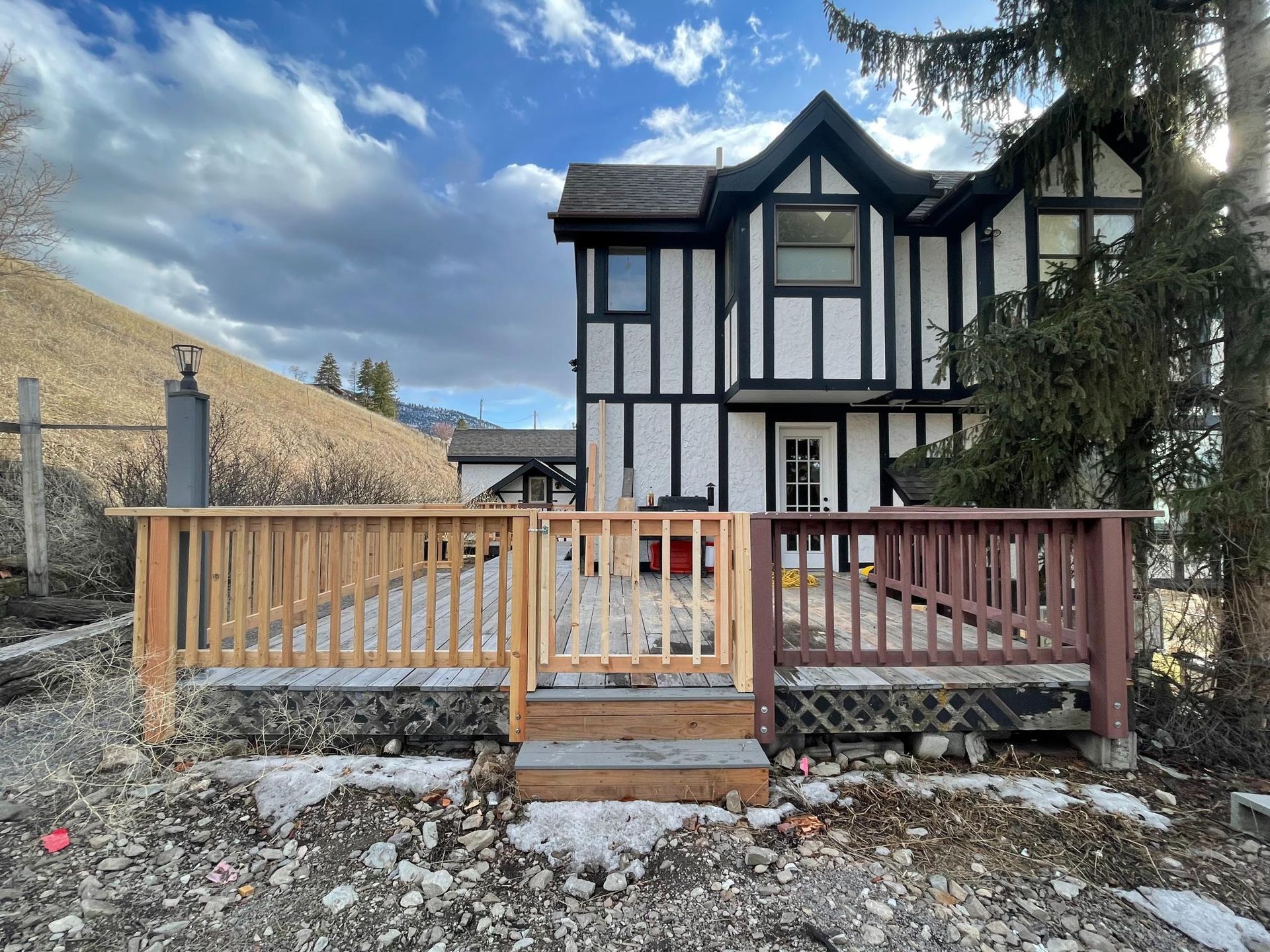 Two-story house with Tudor style facade, a wooden deck, and a yard with snow and a hillside background.