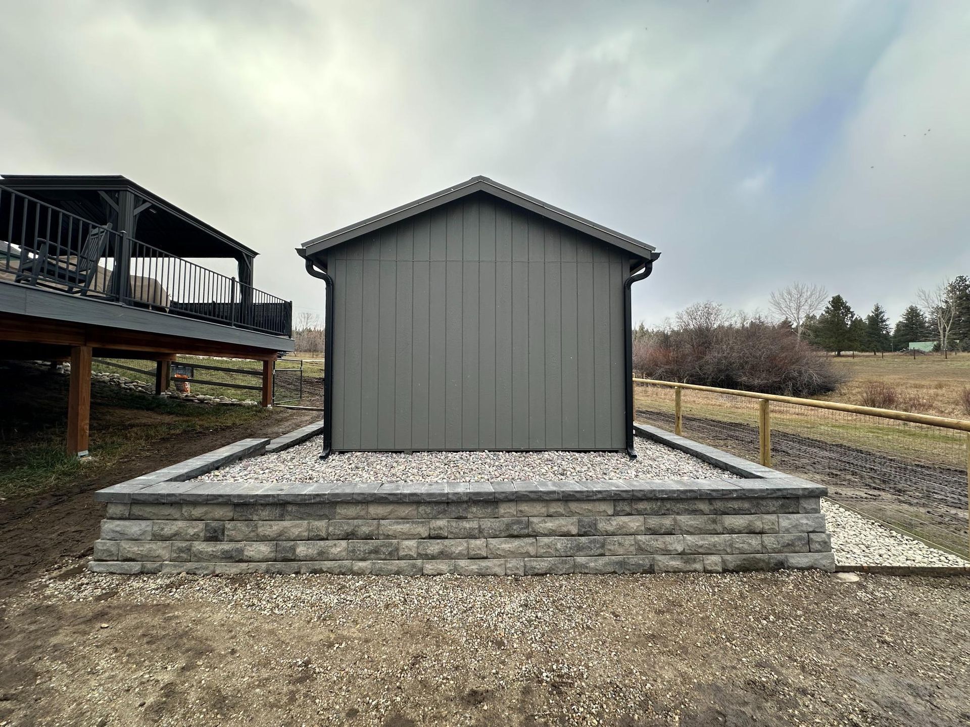 Gray shed on a raised stone platform, next to a deck and fence, under a cloudy sky.