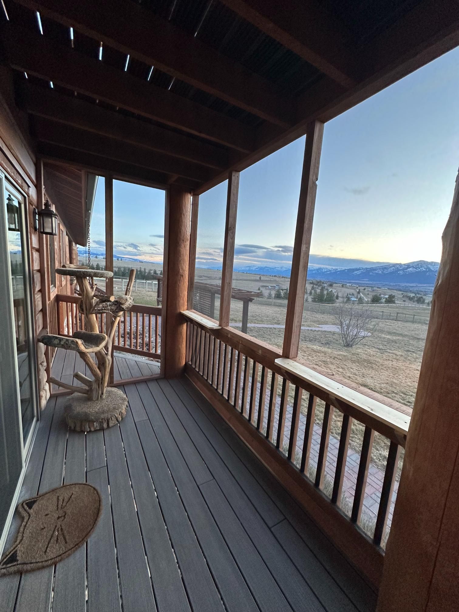 Screened porch of a cabin overlooking a vast landscape. Brown wood, gray deck, cat tree, and a sunset.