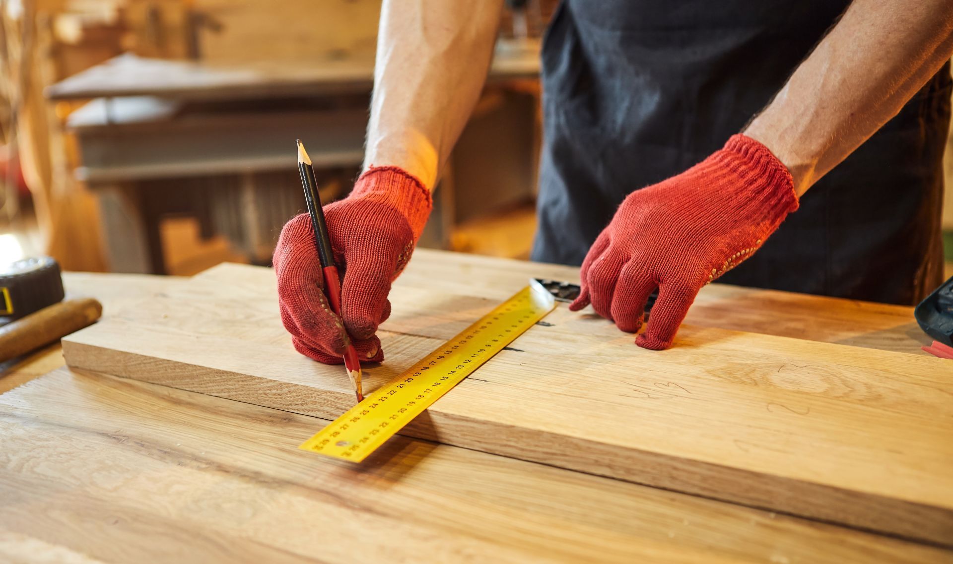 Person in red gloves using a ruler and pencil to mark measurements on a wooden plank at a workshop.