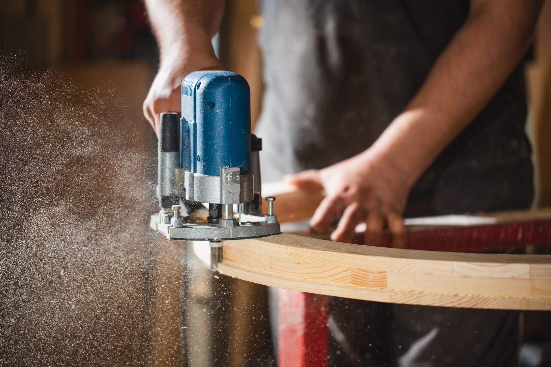 Person using a blue router to shape a curved piece of wood, generating sawdust.