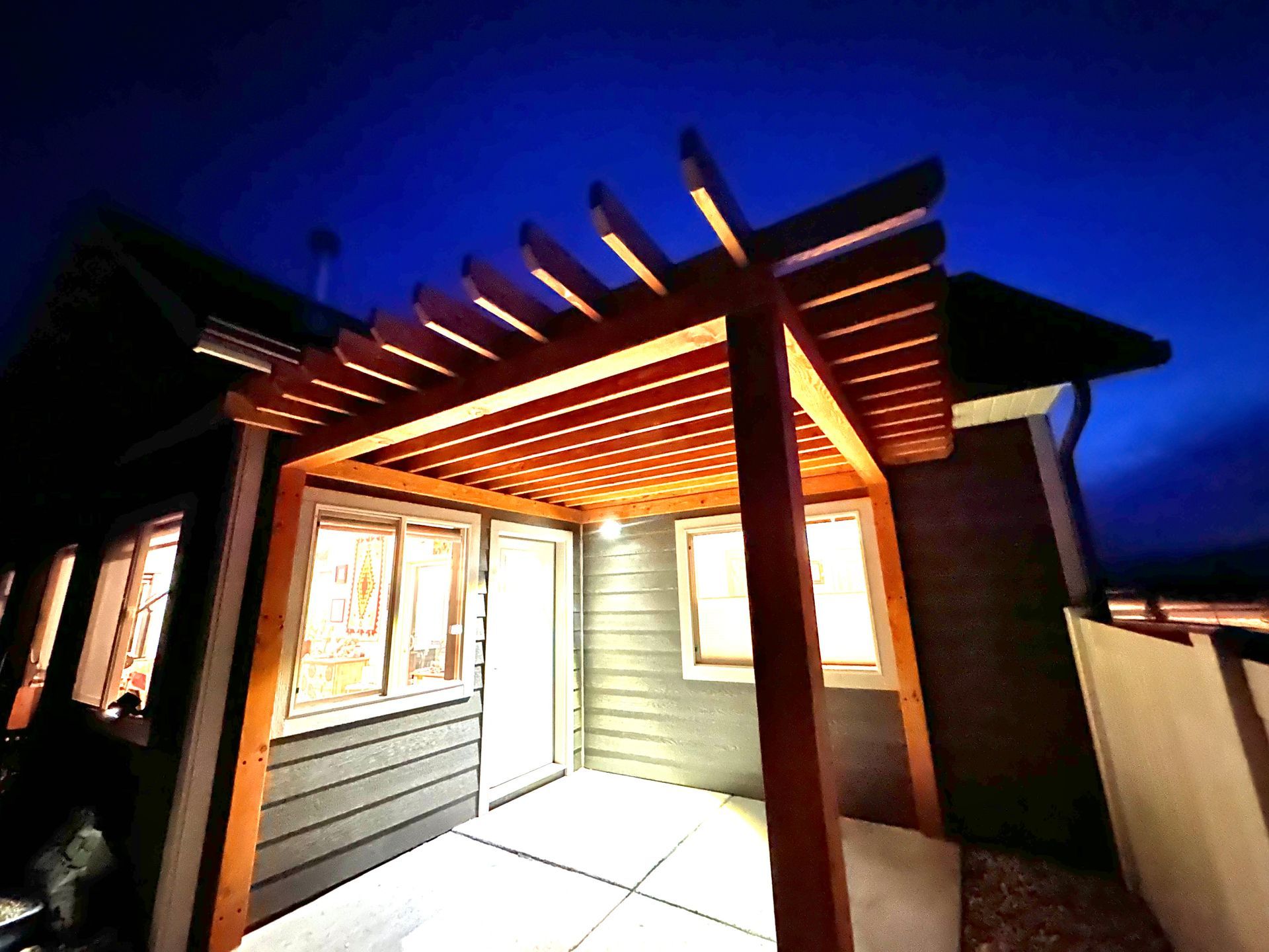 Wooden pergola over a patio, lit up at night with dark blue sky.