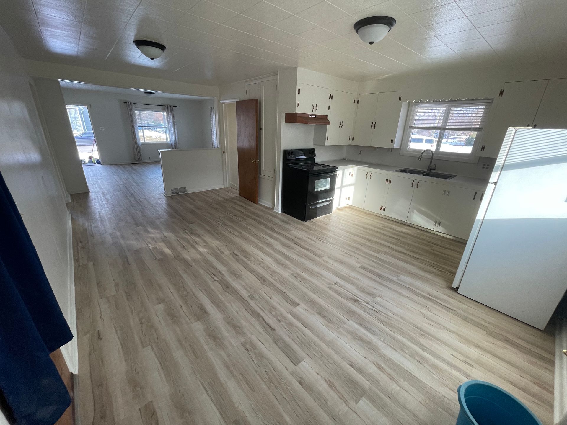 Interior view of a kitchen and living space with new light wood flooring and white walls. A black stove is in the kitchen.