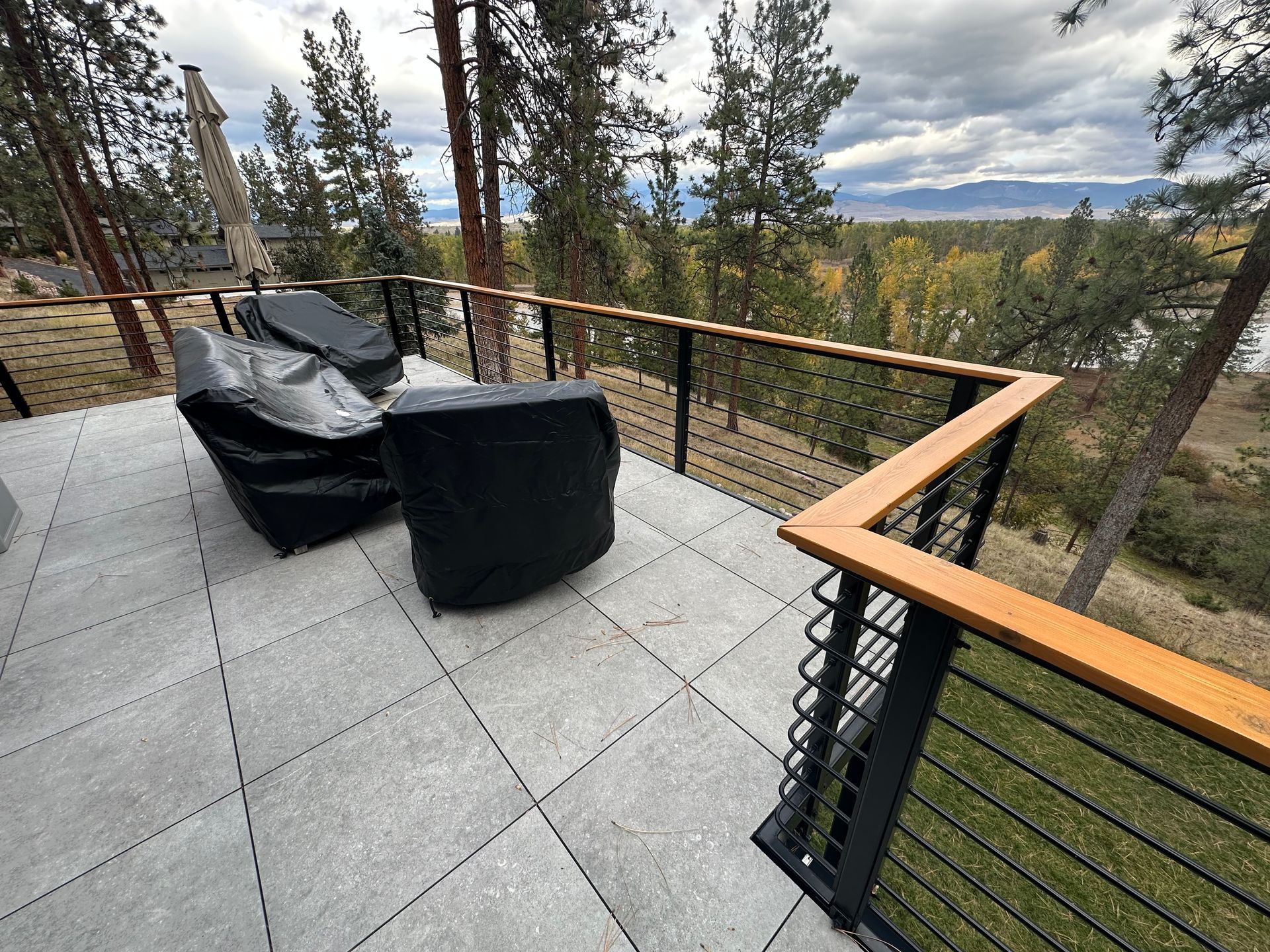 Patio with covered chairs, gray tiles, cable railing, and a scenic view of trees and water.