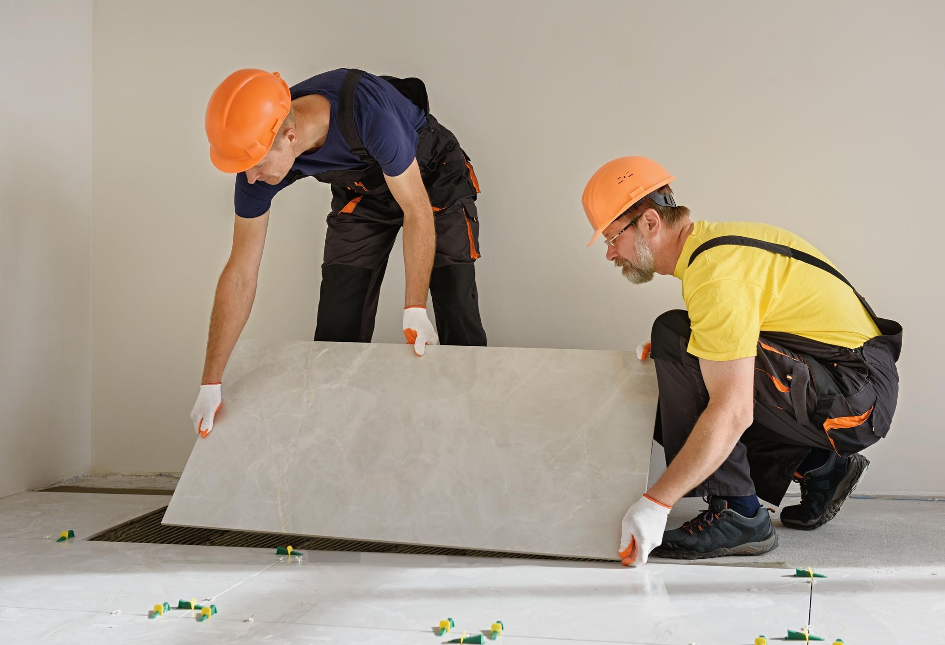 Two workers install a large tile on a floor, wearing hard hats and work clothes in a room.