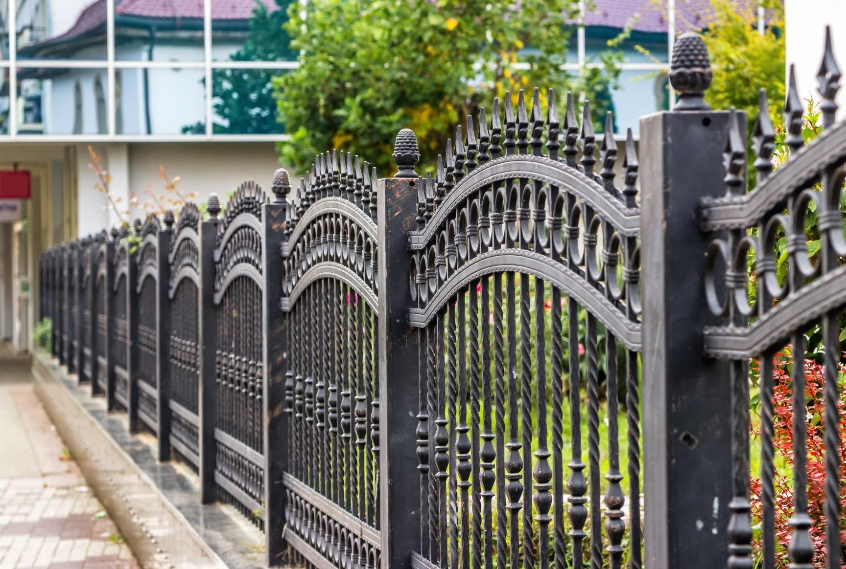 Black ornate metal fence along a sidewalk, with a building in the background.