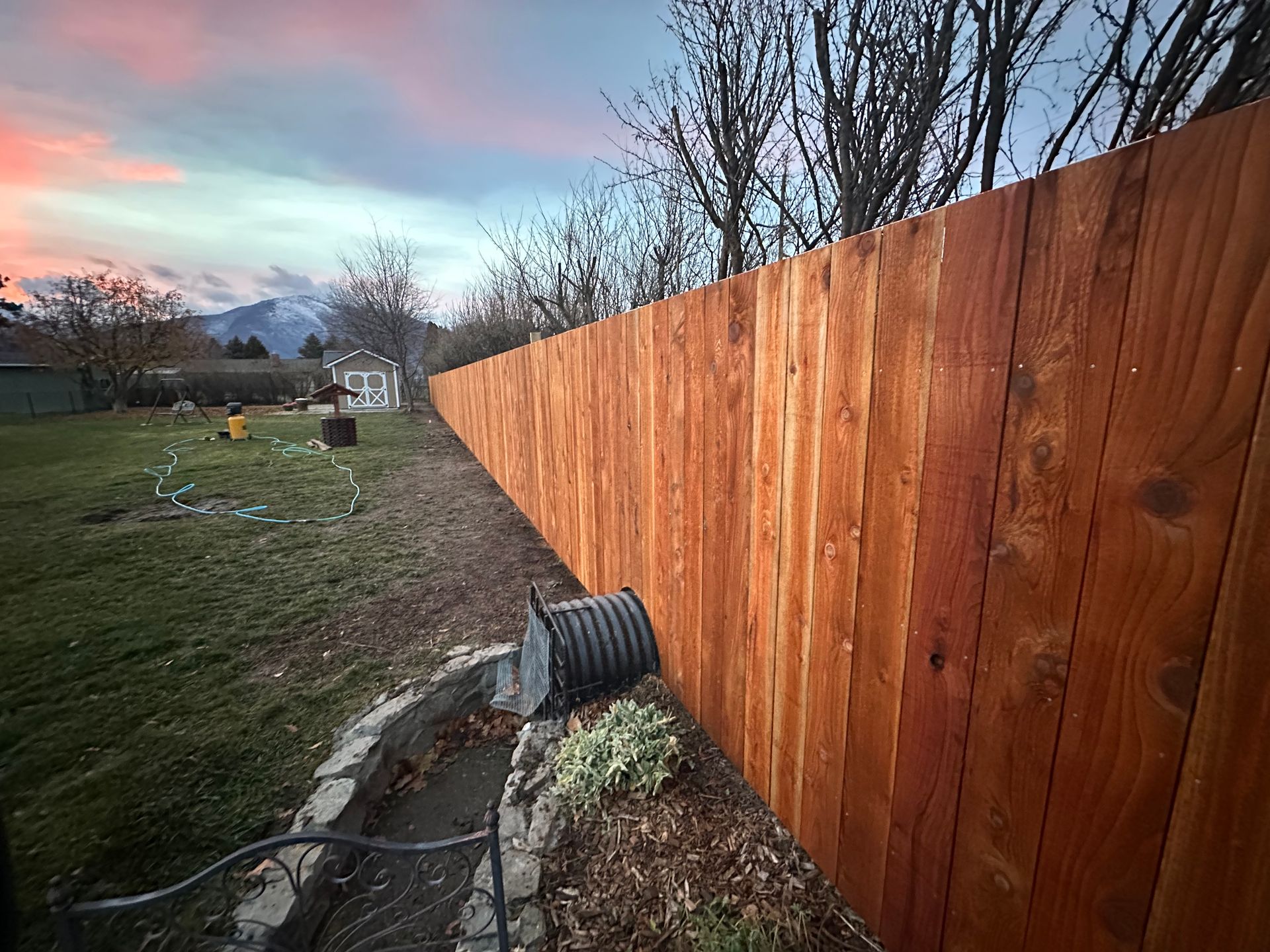 Wooden fence along a grassy yard with a hose, flowers, and sunset sky in the background.
