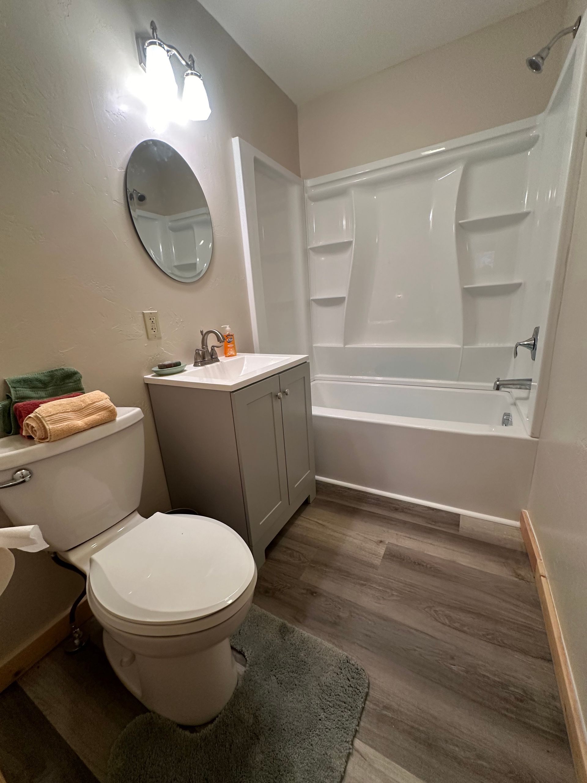 Bathroom with a toilet, vanity, tub/shower, and round mirror. Gray wood-look floor.