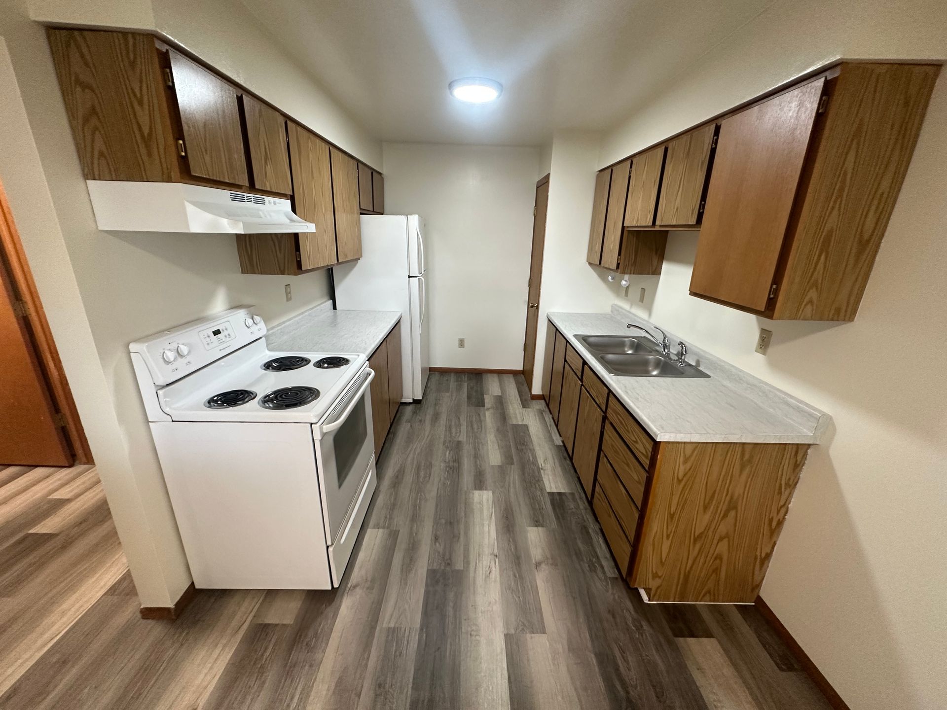A kitchen with wooden cabinets, white appliances, gray flooring, and a window.