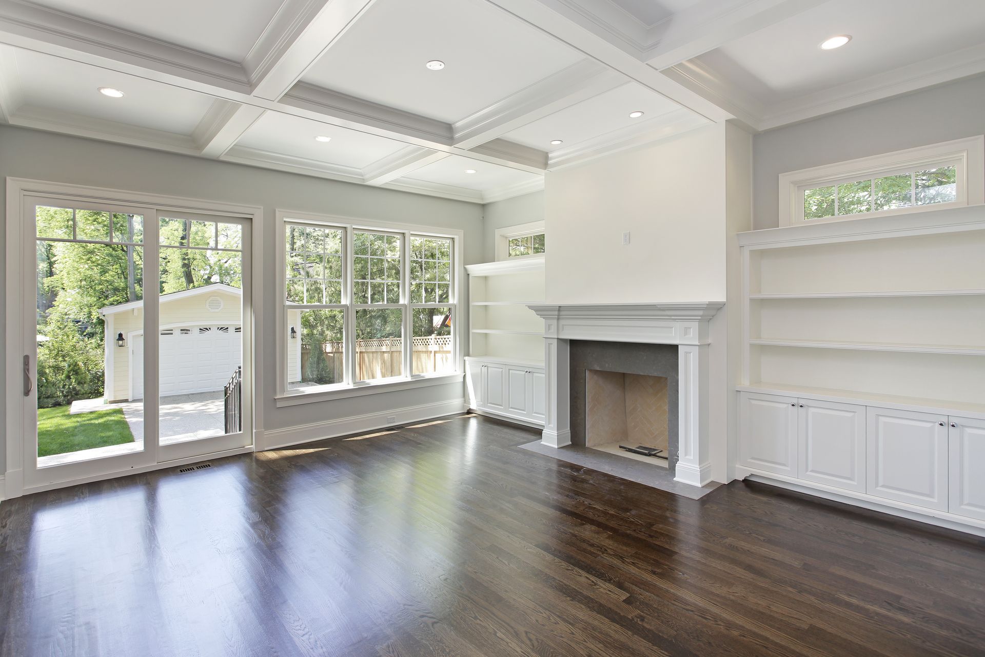Bright empty living room with dark wood floors, white walls, fireplace, built-ins, and large windows overlooking a patio
