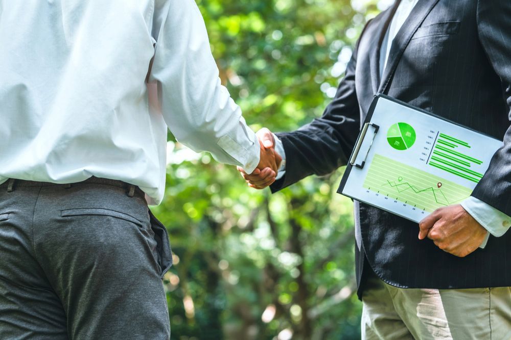 Two men are shaking hands in a park while one of them is holding a clipboard.