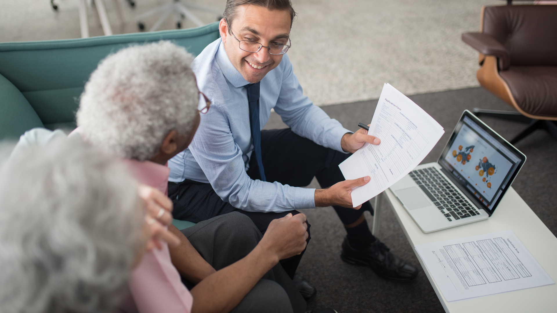 A man is sitting on a couch talking to two elderly people while looking at a laptop.