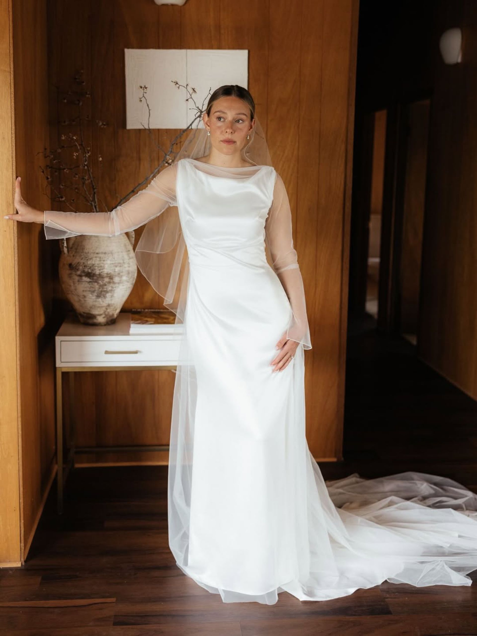 Bride in white gown with sheer sleeves and veil, poses indoors near wooden wall.