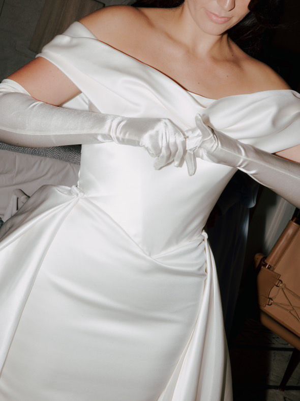 Woman in a white off-the-shoulder wedding gown, adjusting satin gloves.