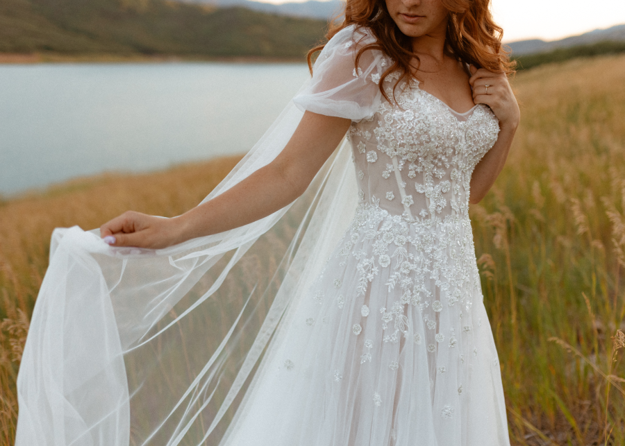 Woman in a white wedding dress with floral embellishments and a sheer cape, outdoors near a lake.