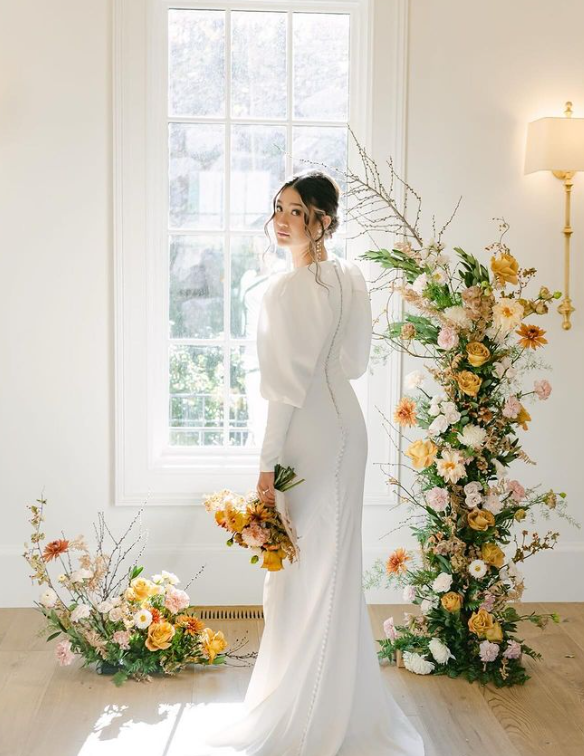 Woman in white wedding dress with floral arrangement by a window.