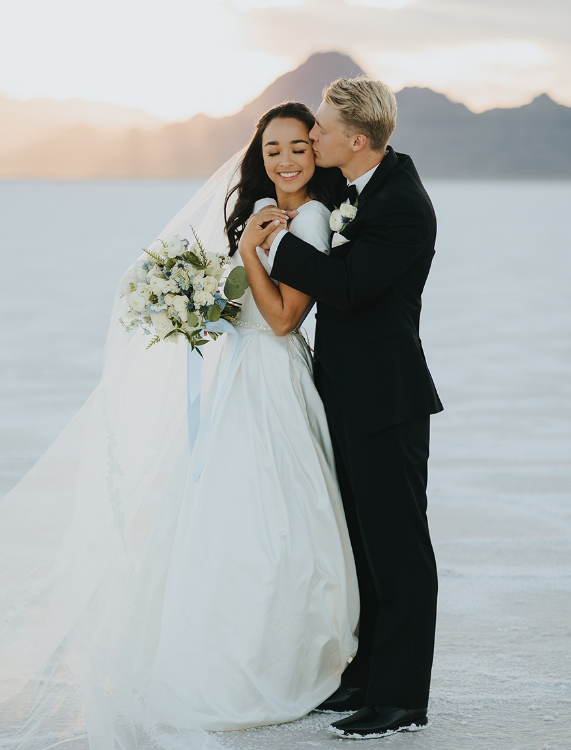 Bride and groom embrace, he kisses her cheek. They stand on a salt flat with mountains in the background.
