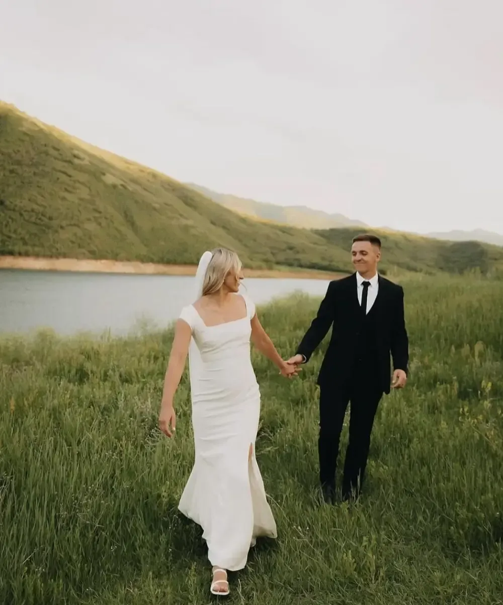 Bride and groom hold hands walking in a field. Bride in white dress, groom in black suit, lake and mountains in background.