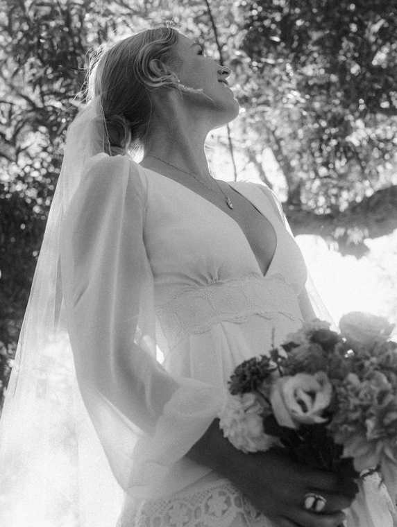 Bride in white dress holding flowers, looking up, standing in a sunlit garden.