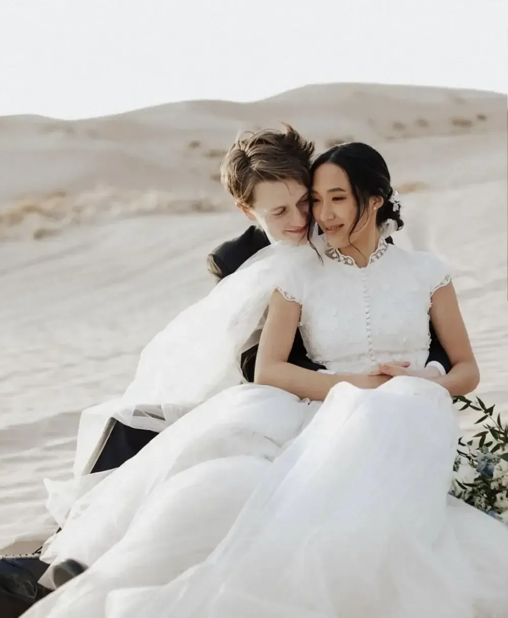 Couple in wedding attire embrace on sand dune, soft lighting.