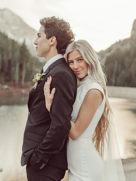 Bride in white dress leans on groom's shoulder, smiling, near a lake. Mountains in background.