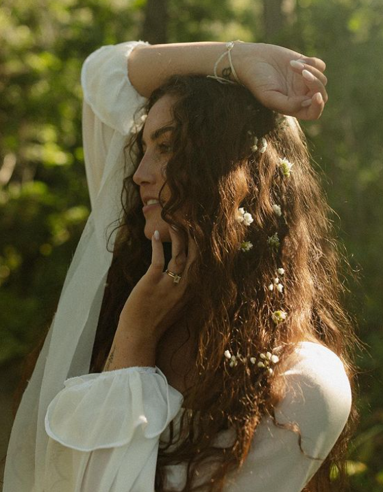 Woman with curly hair adorned with flowers, wearing a white blouse, looking away with hand raised. Outdoors.