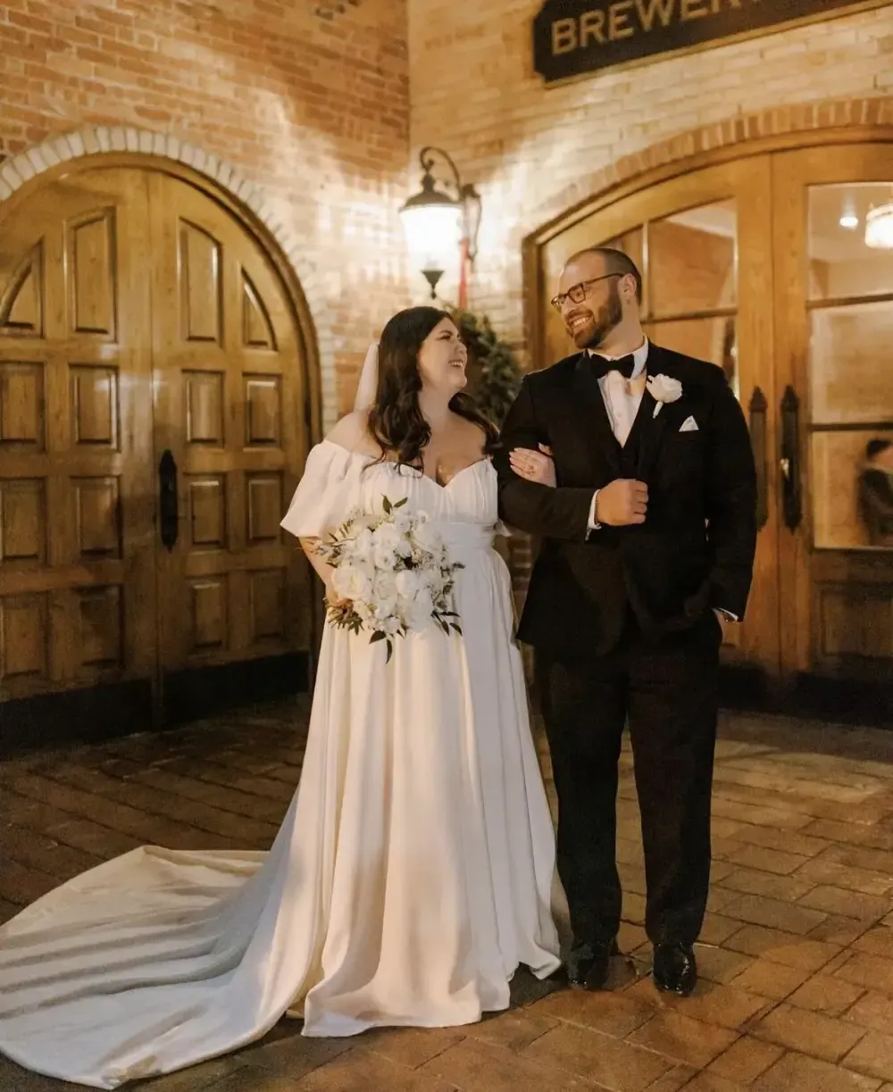 Bride and groom pose outside brewery doors. The bride wears a white gown, and the groom a black tuxedo.