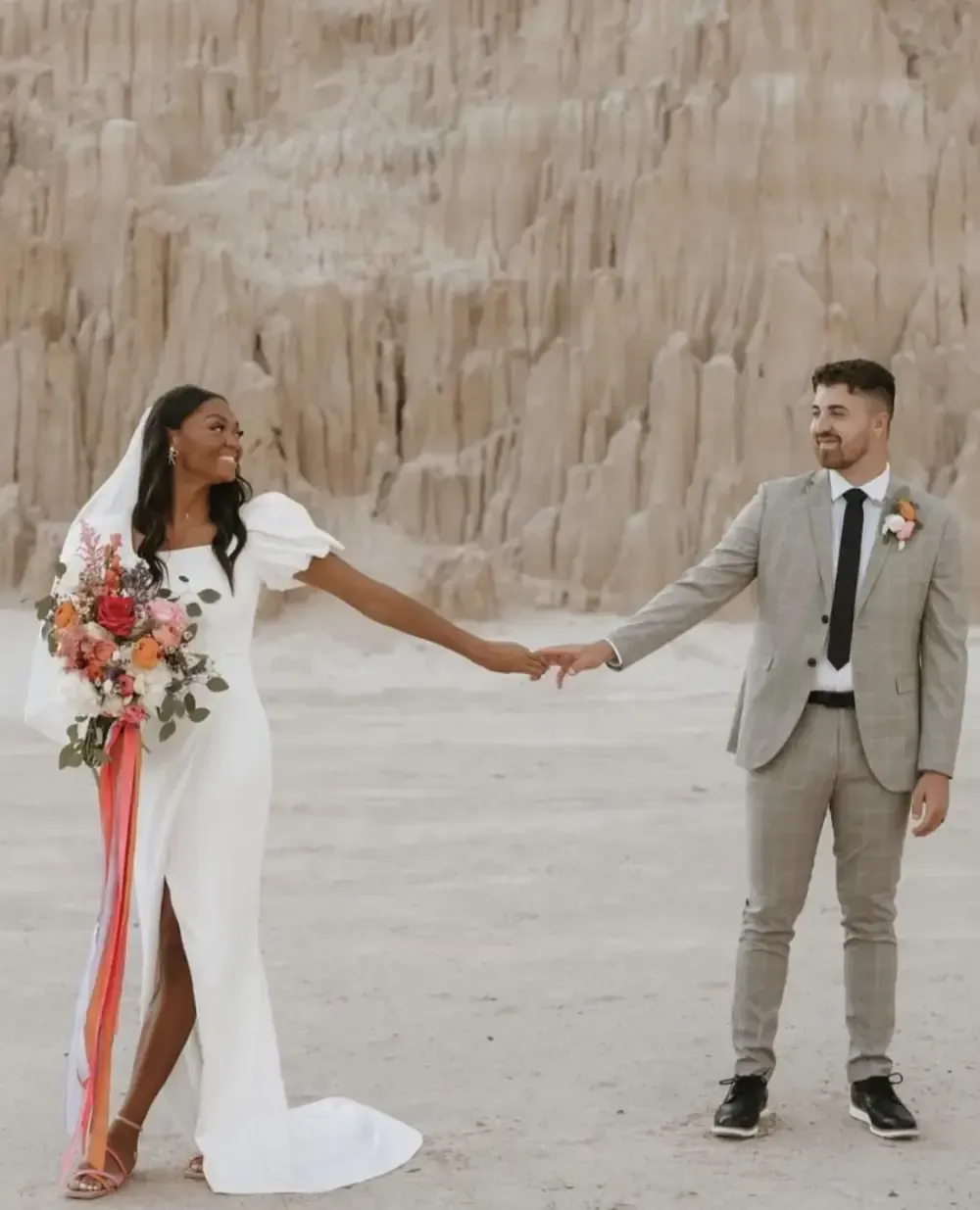 Wedding couple holding hands; woman in white dress, man in gray suit, desert backdrop.