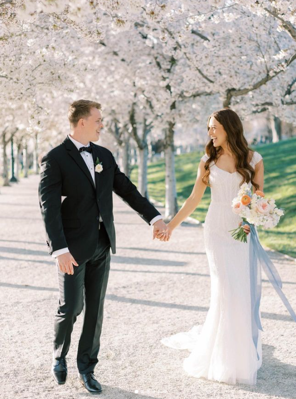 Couple in wedding attire hold hands and walk on path lined with blossoming trees.
