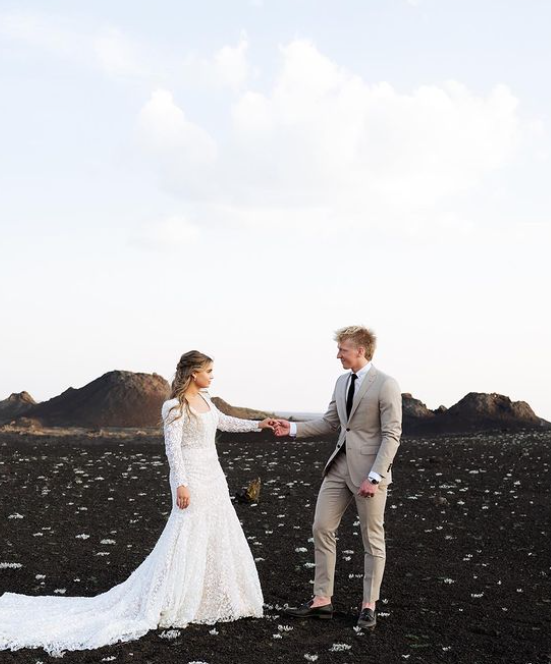 Bride and groom holding hands, standing on black volcanic landscape, blue sky.