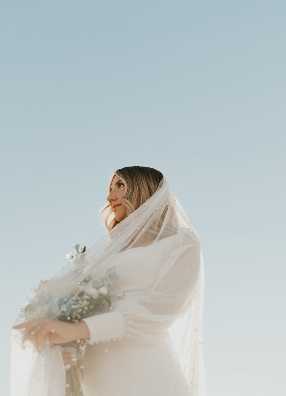 Bride in a white gown and veil, holding flowers, against a clear blue sky.