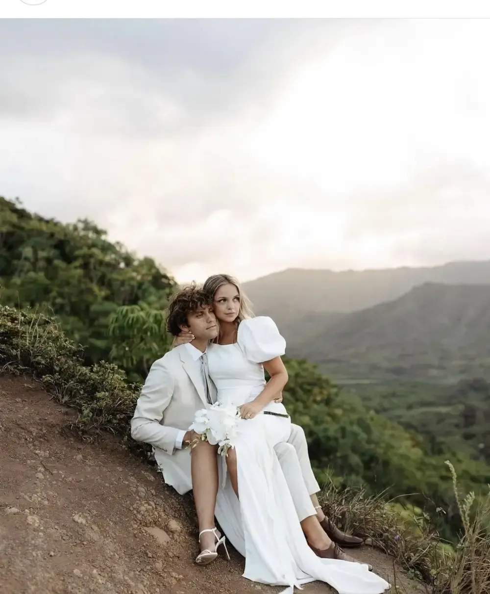 Couple in wedding attire sitting on a hillside, overlooking a valley at sunset.