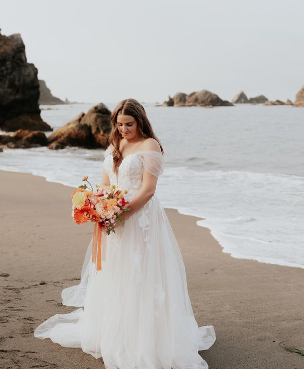 Woman in wedding dress on beach, holding bouquet, looking down. Ocean waves in the background.